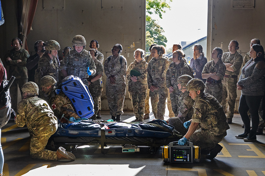 A TMW instructor, in camouflage uniform and combat helmet, gestures with a blue-gloved hand while teaching a class inside a hangar bay. A group of service members stands in a semi-circle listening intently, while other medics demonstrate the use of medical equipment around a patient simulation bag and stretcher on the floor.
